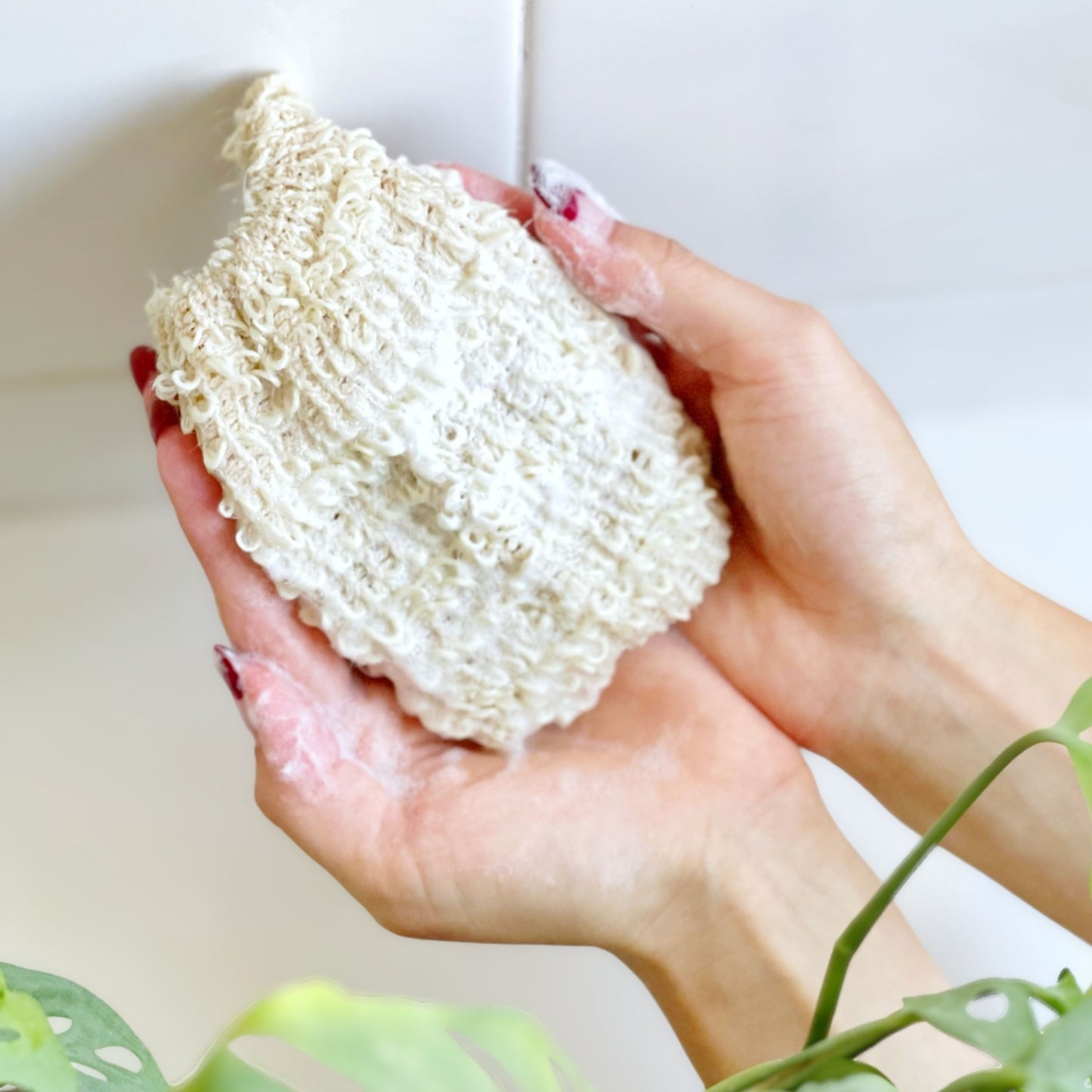 A textured sisal soap saver pouch foaming with soapy lather being held in a person's hands in a bright shower.