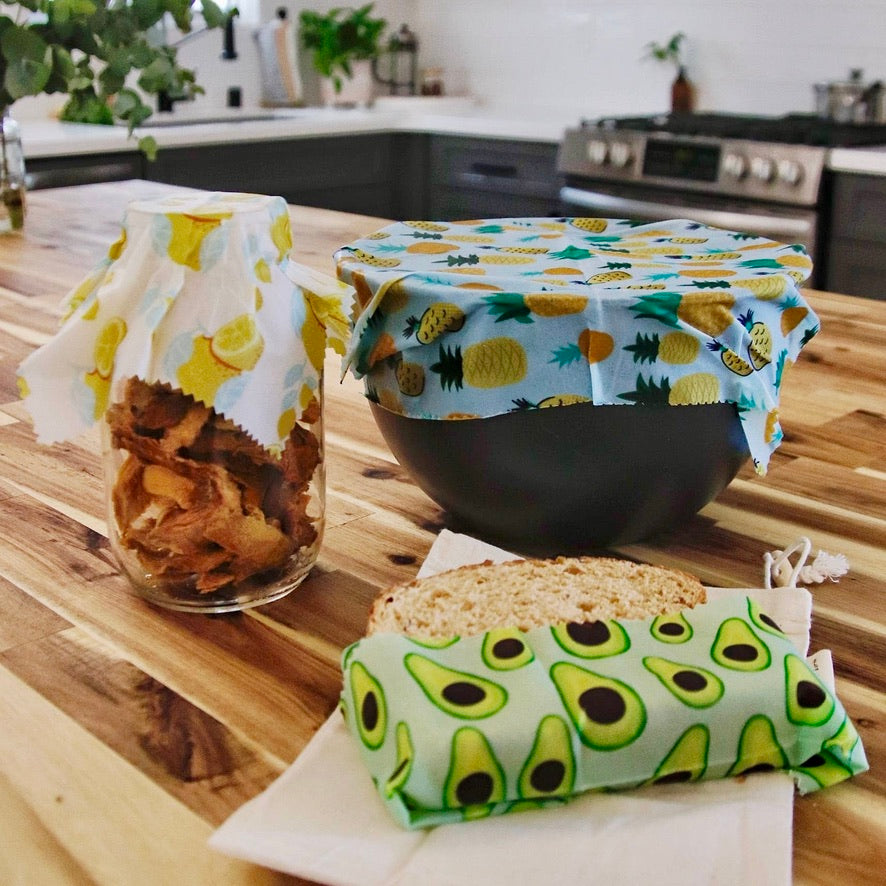 Reusable wax wraps covering a glass jar and bowl of snacks on a kitchen counter, showing pineapple and avocado prints in use.