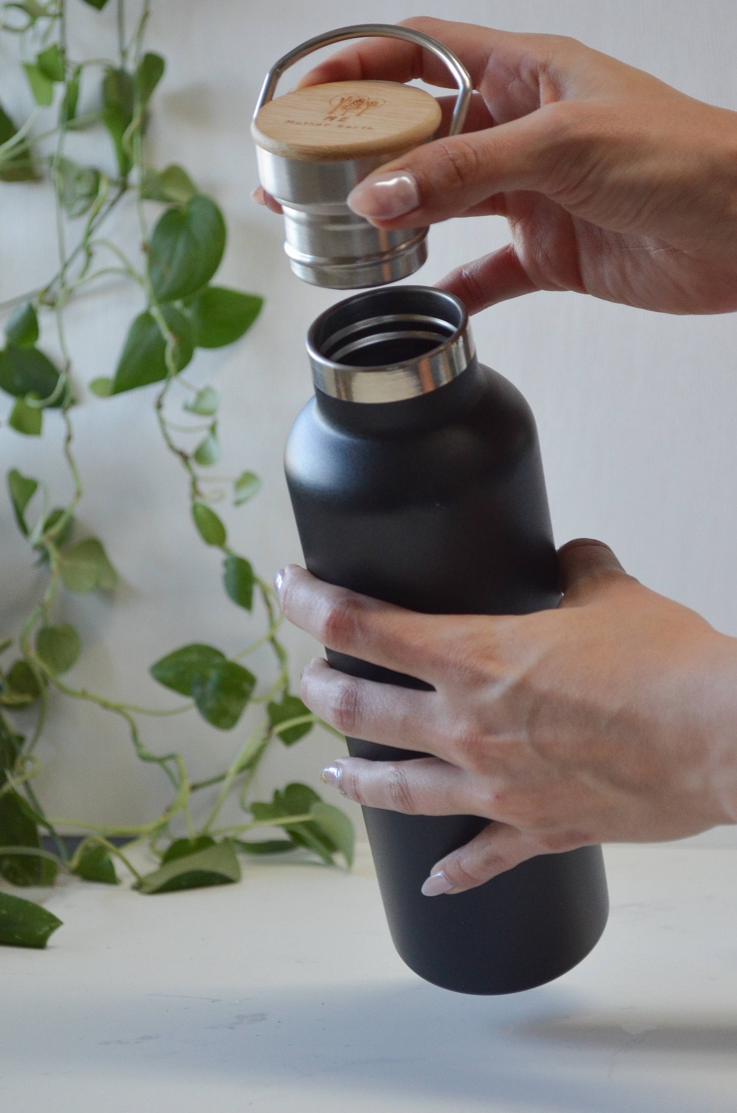 Hands unscrewing the bamboo lid of a matte black stainless steel water bottle to reveal the metal threads.