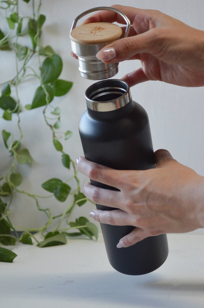 Hands unscrewing the bamboo lid of a matte black stainless steel water bottle to reveal the metal threads.
