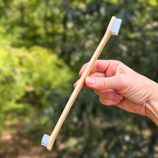 Hand holding a double-ended bamboo dog toothbrush with a large brush head on one end and a small one on the other, against green foliage.