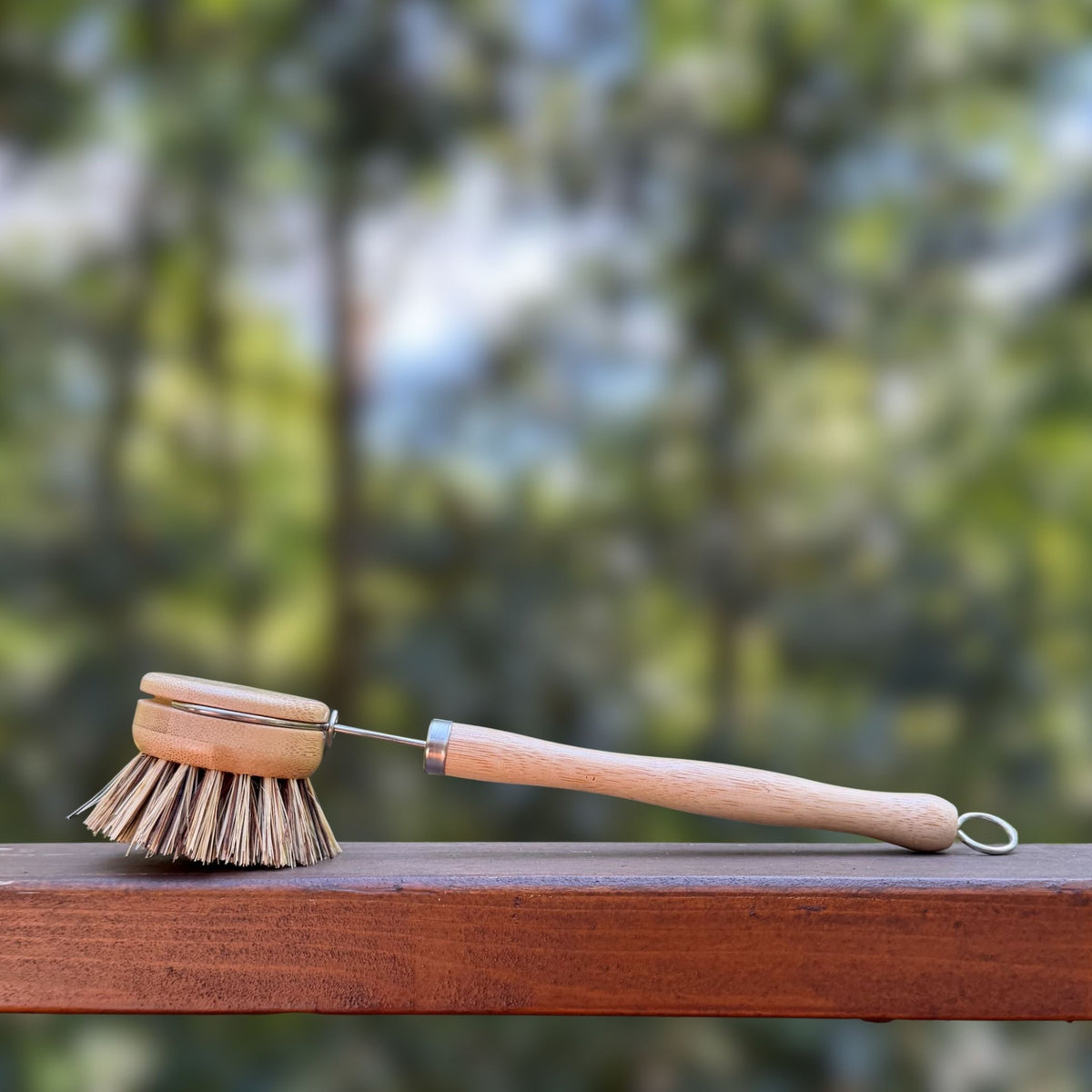A wooden sisal and palm bristle cleaning brush with a light wood handle rests on a wooden railing