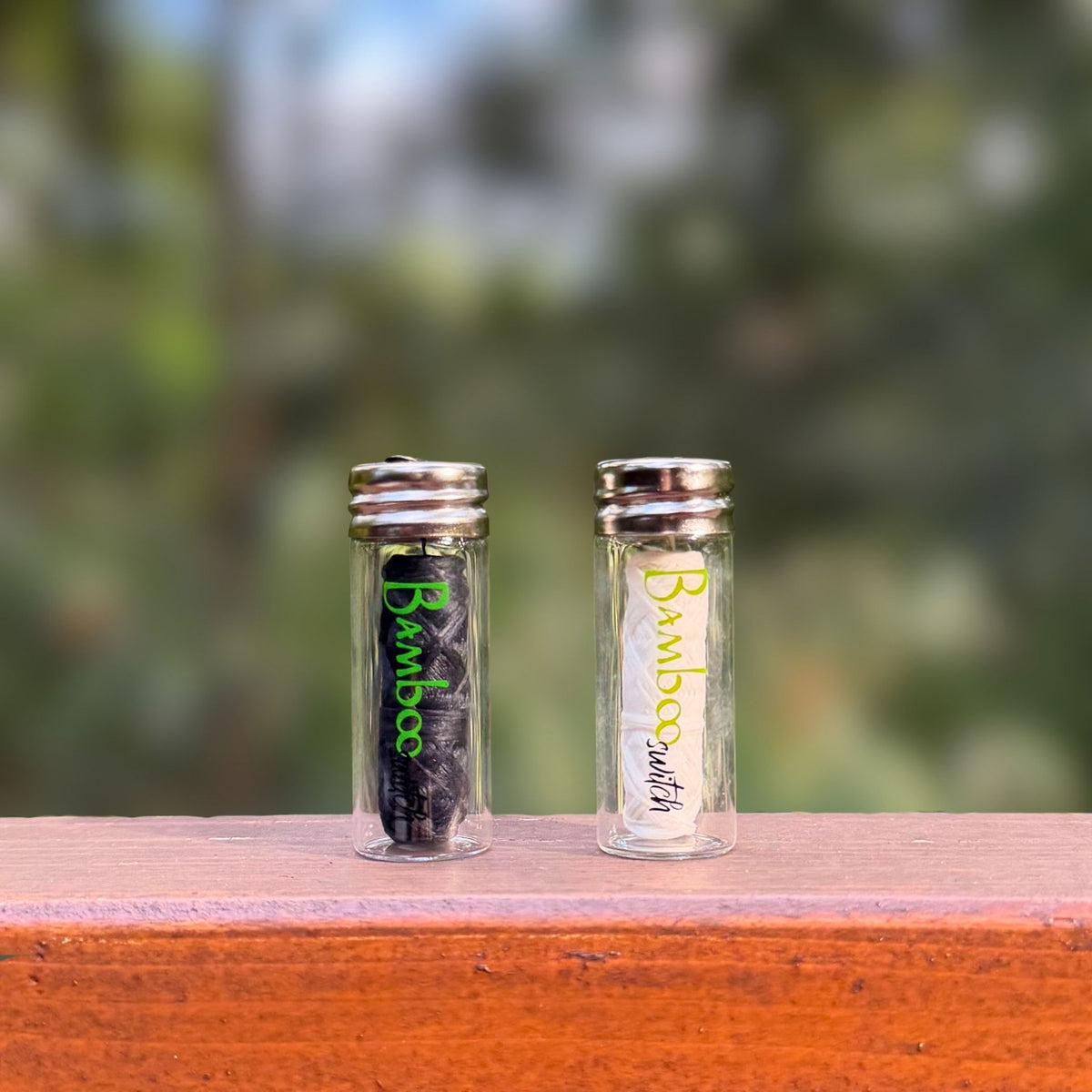 Charcoal and white Bamboo Switch bamboo floss in refillable glass jars side by side on a wooden railing