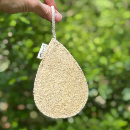 Natural loofah sponge with cotton hanging loop held against green foliage background