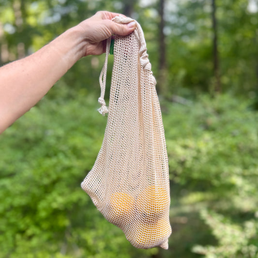Cotton mesh produce bag held up by its drawstring, hanging empty against a wooded backdrop