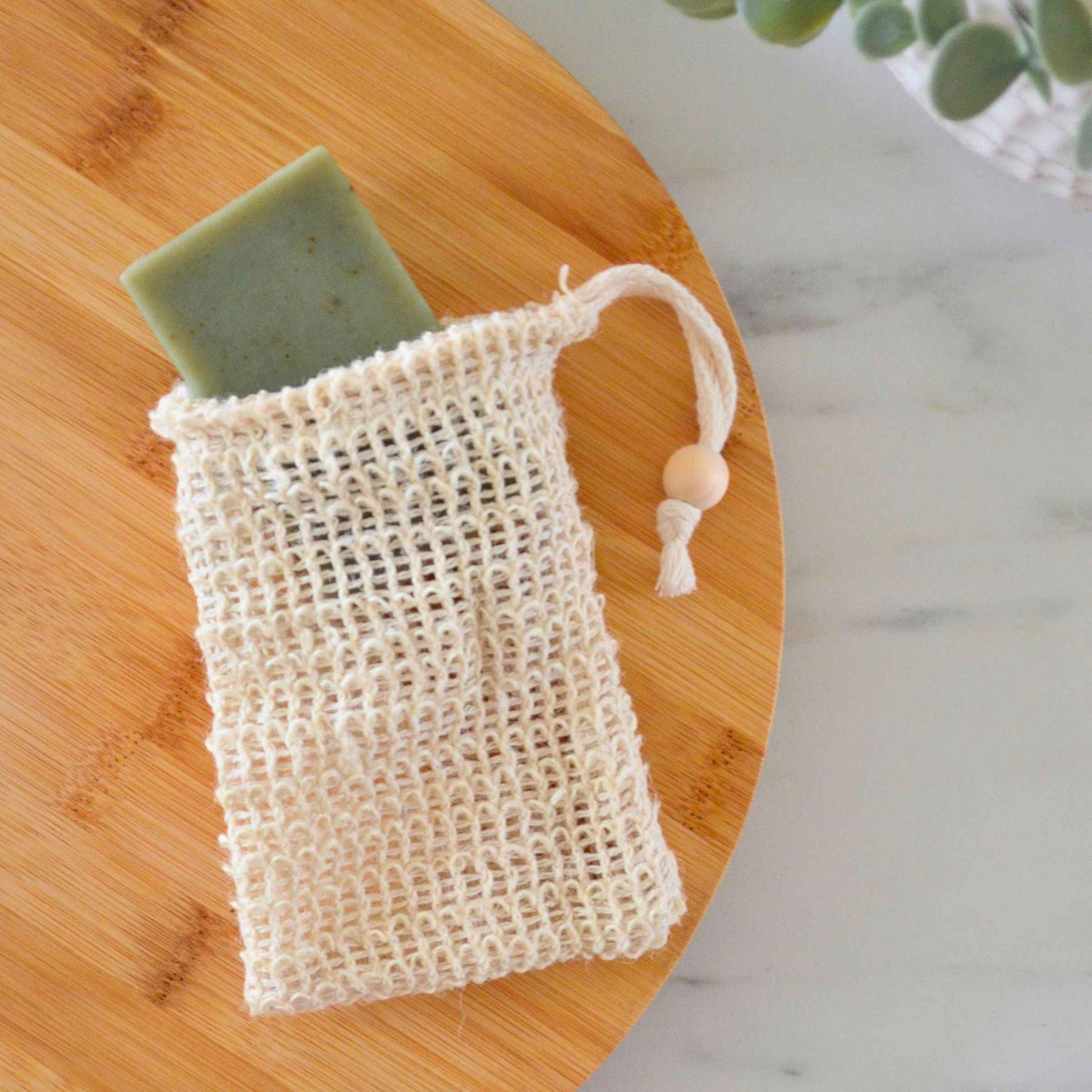 Green natural bar soap sliding into a textured sisal soap saver pouch on a round bamboo tray.