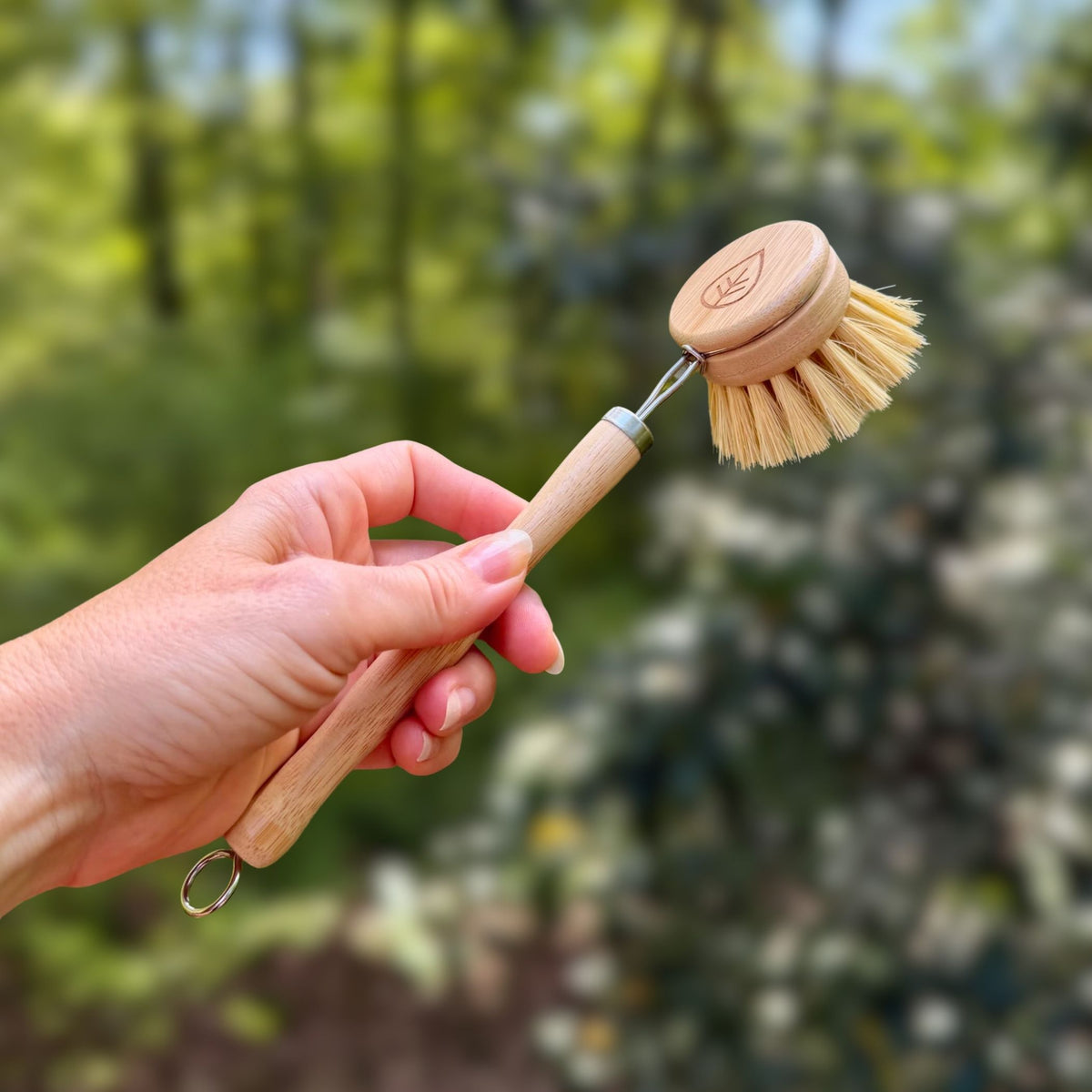 Hand holding a sisal dish brush with bamboo head and metal hanging loop against green trees