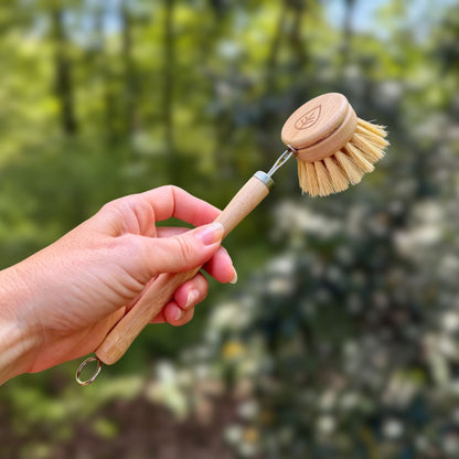Hand holding a sisal dish brush with bamboo head and metal hanging loop against green trees