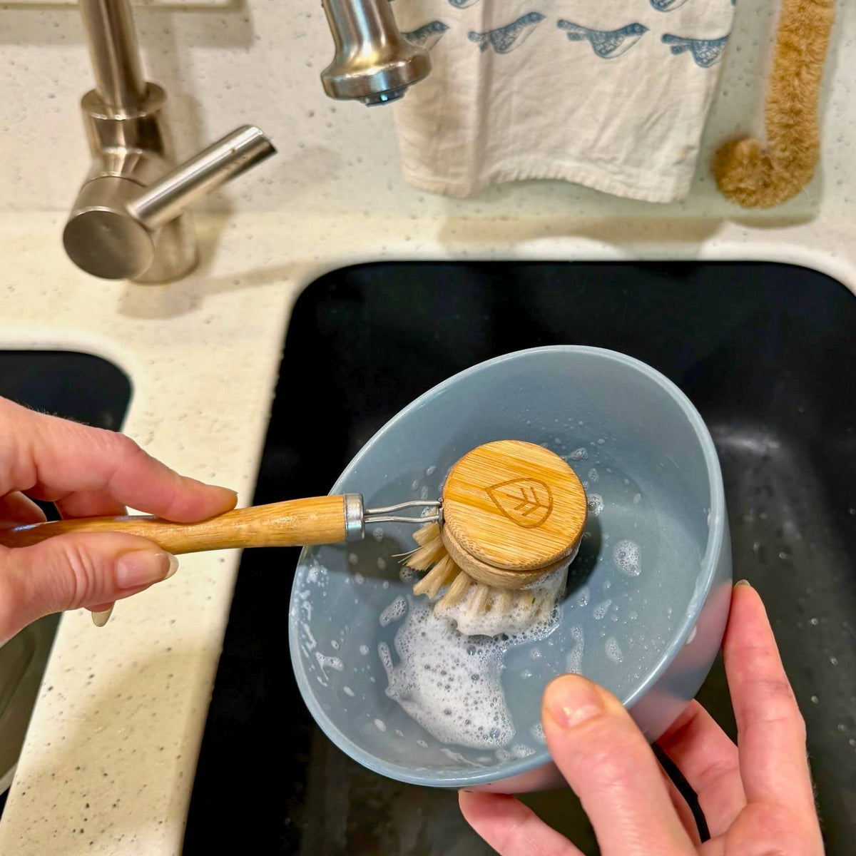 Hand scrubbing a soapy blue bowl with a sisal bamboo dish brush over a black kitchen sink