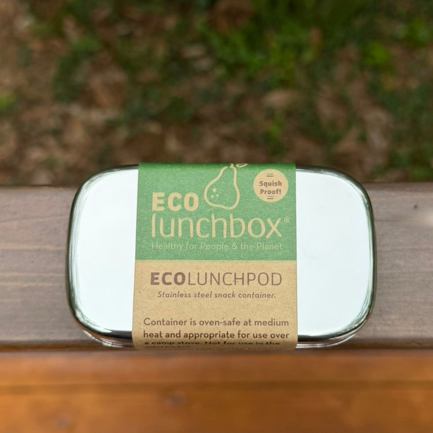 ECOlunchbox stainless steel snack container with paper packaging on a wooden ledge.