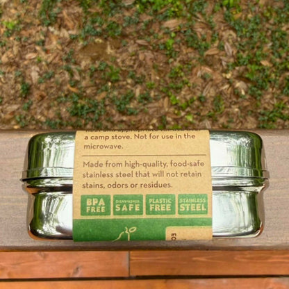 ECOlunchbox stainless steel snack container on its side with a paper product label on a wooden ledge.