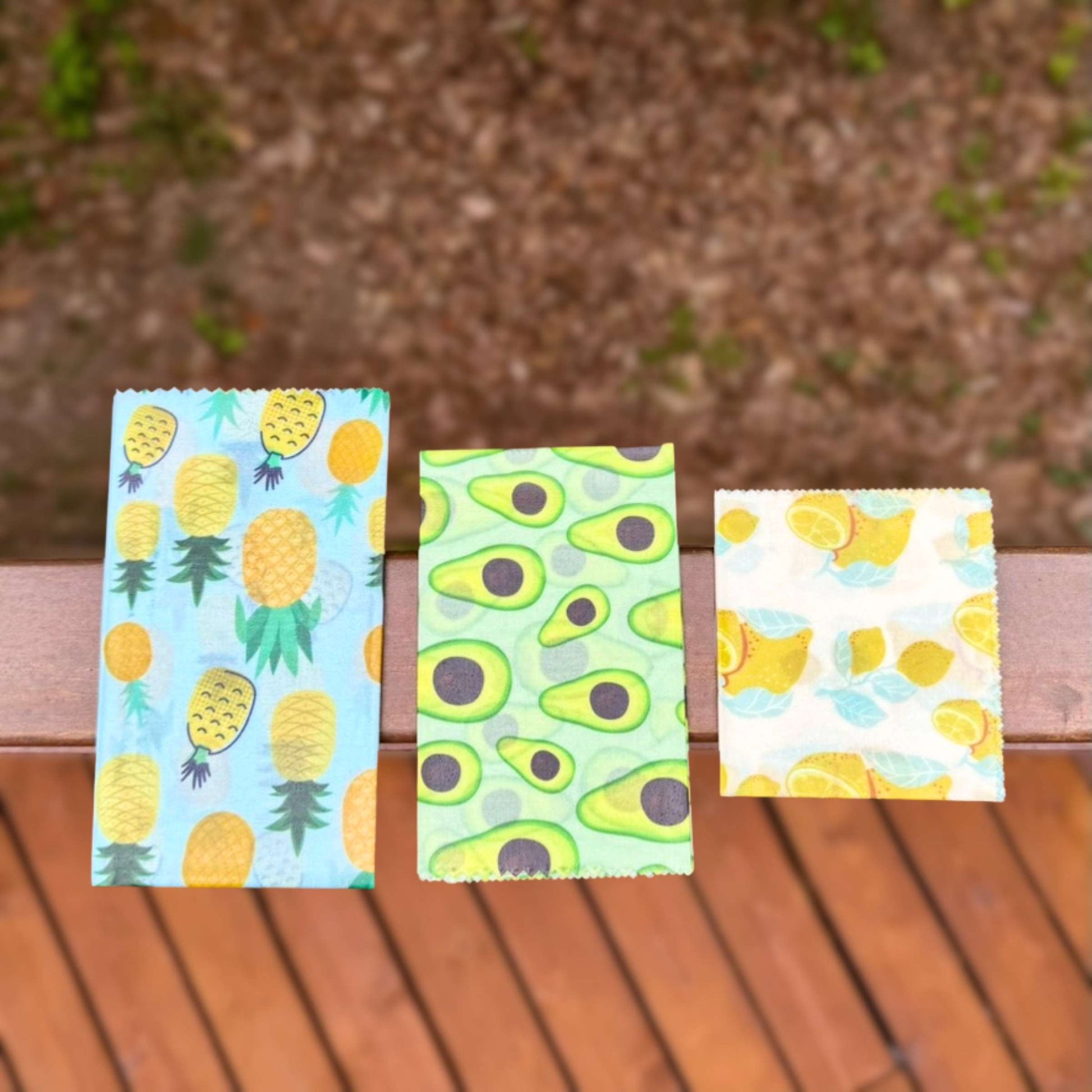Three reusable wax food wraps in small, medium, and large sizes with pineapple, avocado, and lemon prints, laid on a wood surface.
