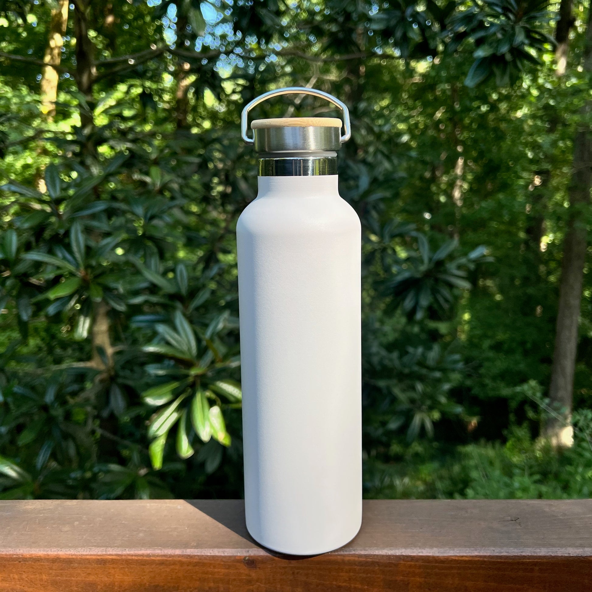 Insulated matte white stainless steel water bottle with a bamboo lid and metal carrying loop, sitting outdoors.