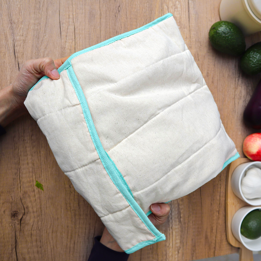 White quilted produce bag with turquoise trim held by a hand on a wooden surface with fruits and kitchen items.
